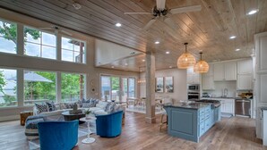 A spacious living area with a large sofa, blue armchairs, and a dining table. There is a kitchen with white cabinets and a blue island. - Save 15% Off 4 Night Stay! Amazing Waterfall/Lake Views! Pool, Dock! (Vinita)