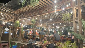 Zona pranzo all'aperto con travi in legno e luci a tendina, immersa nel verde e con tavoli e sedie.