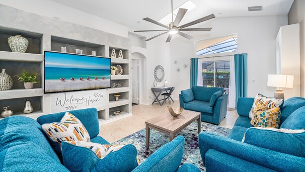 A modern living room with a large flat-screen TV, blue sofas, a coffee table, and a ceiling fan. - Pool Home with Spa & Games Room in Gated Community Near WDW (Davenport)