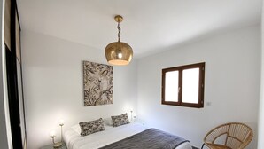 A bedroom with a bed, a hanging light fixture, a window, a wall-mounted artwork, and a wicker chair. - Residence La Listrella apt 2 (Monticello)