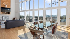A spacious living room with a large glass table and chairs, a grey sofa, and a city view through large windows. - Nashville Skyline Views - 3BR/3BA, 5 Mins to Downtown (Nashville)