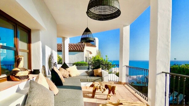 Un balcon avec vue sur la mer, un canapé avec des coussins, un luminaire suspendu et une table basse en bois.