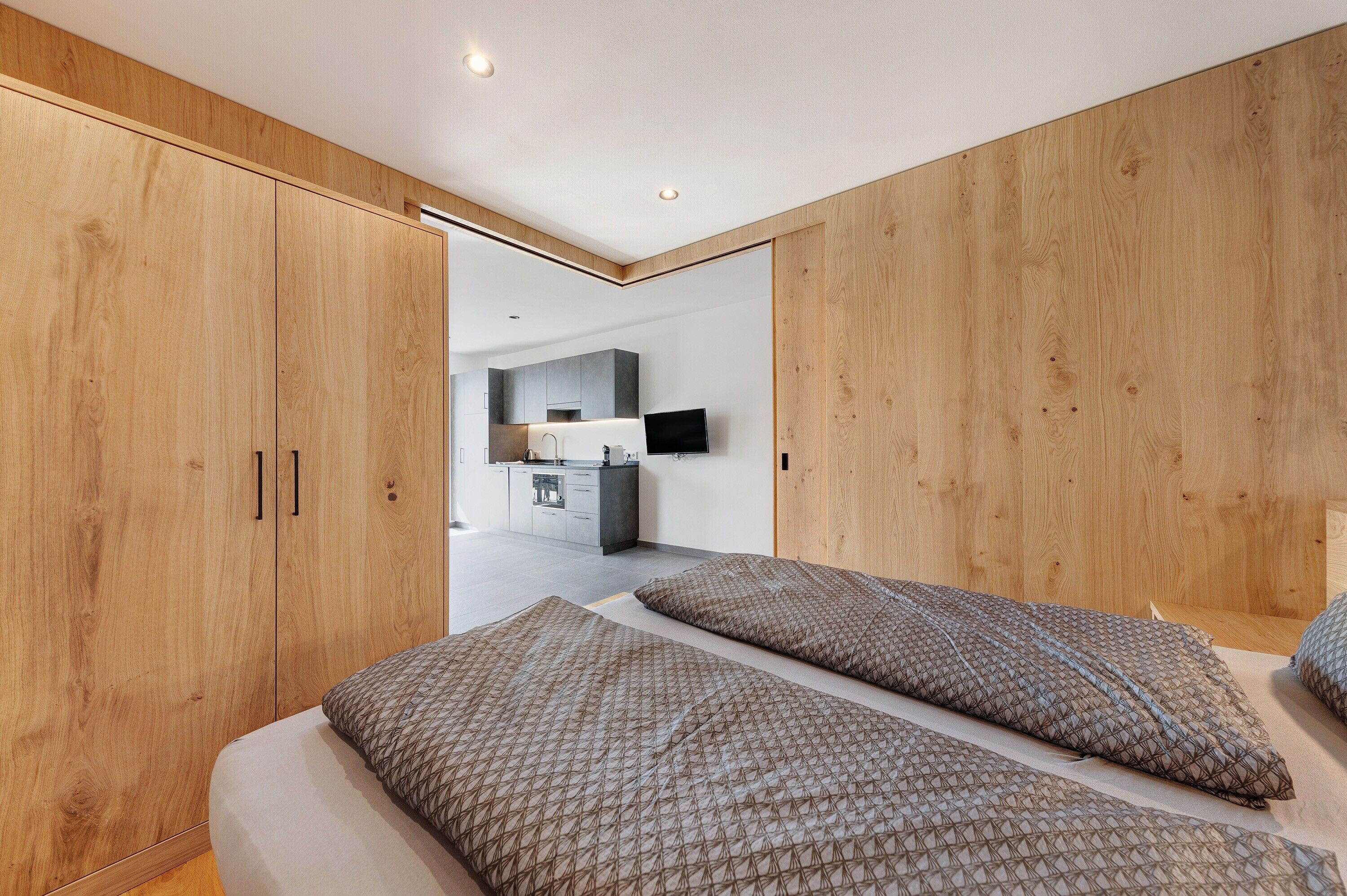 A modern bedroom with a large wardrobe, a bed with patterned bedding, and a view into a kitchen area.