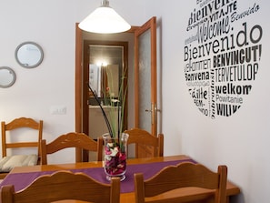 A dining area with a table, chairs, a vase with flowers, and a wall with a welcome sign in multiple languages.
