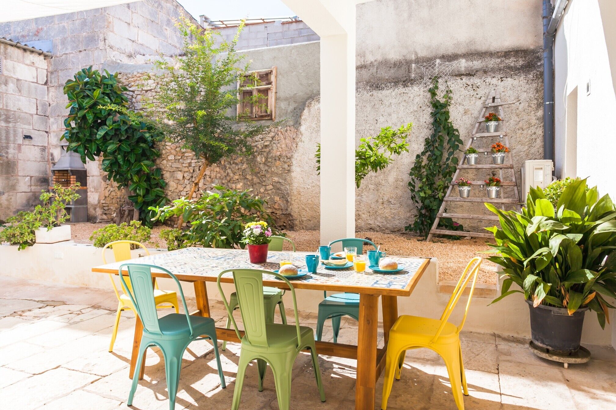Un patio al aire libre con una mesa de madera y sillas coloridas, rodeado de plantas en macetas y un muro de piedra.