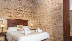 A bedroom with a stone wall, a wooden bed frame, a bedside table with a lamp, and a nightstand. - Holiday Home Sa Lluna Blava with Pool & Wi-Fi (Santa Margalida)
