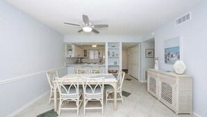 A dining area with a white table and chairs, a ceiling fan, and a kitchen in the background. - Runaway Bay Resort by BBG (Bradenton Beach)