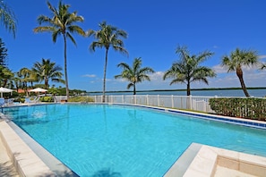 A clear blue swimming pool with white poolside chairs and palm trees, surrounded by a white fence and a view of a lake. - Harbor Villa Club 312 (Longboat Key)