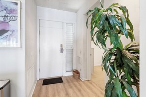 A hallway with a white door, a wooden floor, a black doormat, a woven basket, and a large green plant. - Harbor Villa Club 312 (Longboat Key)