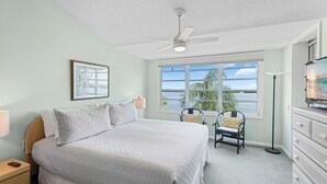Un dormitorio con cama, ventilador de techo, ventana con vistas, una silla, lámpara y cómoda.