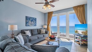Ein Wohnzimmer mit grauem Sofa, Couchtisch, Deckenventilator und großem Fenster mit Vorhängen.