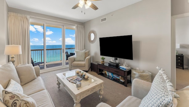 A living room with a large TV, a ceiling fan, a sofa, a coffee table, and a view of the ocean through a sliding door. - Sunrise Resort by BBG (Panama City Beach)