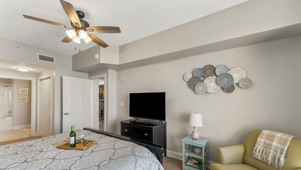 A bedroom with a bed, a ceiling fan, a TV, a wall decor, a side table, and a chair. - Sunrise Resort by BBG (Panama City Beach)