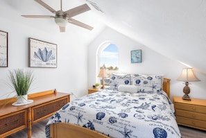 A bedroom with a sloped ceiling, a bed with ocean-themed bedding, a desk with a plant, and a view of the sky through a window.