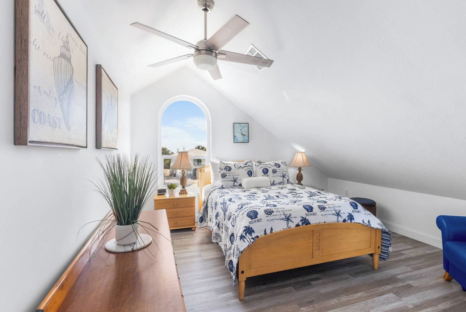 A bedroom with a wooden bed, a ceiling fan, a nightstand, a lamp, a potted plant, and framed artwork on the wall.