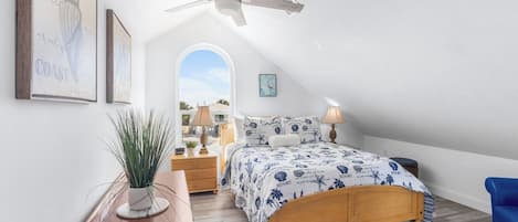 A bedroom with a wooden bed, a ceiling fan, a nightstand, a lamp, a potted plant, and framed artwork on the wall.