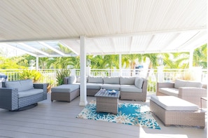 Een terras buiten met rieten meubels, een kamerkleed met patroon en een witte overkapping.