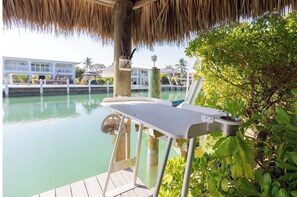 A wooden deck with a table and chair, overlooking a calm body of water, with a thatched roof overhead and lush greenery in the background.