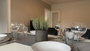 Una mesa de comedor dispuesta con platos, fuentes y copas de vino blancos, y una planta en maceta en el centro.