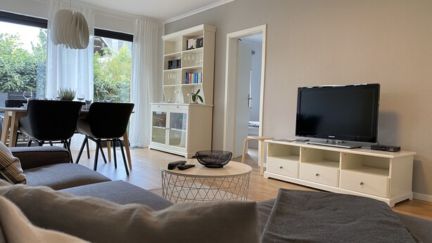 Una sala de estar moderna con sofá, televisor, estantería, comedor y vistas a zonas verdes.