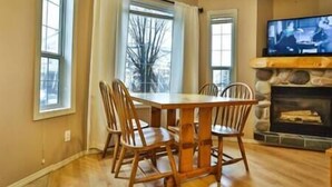 A dining area with a wooden table and chairs, large windows, and a stone fireplace. - 2 BDR Townhouse / Mountain Views / Patio (Fernie)