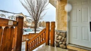 A wooden staircase leading to a white door, with a stone wall and a clear view of the street and houses in the background. - 2 BDR Townhouse / Mountain Views / Patio (Fernie)