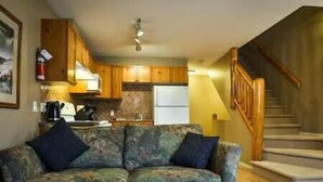 A living room with a sofa, a coffee table, wooden cabinets, a kitchen area, and a staircase. - 2 BDR Townhouse / Mountain Views / Patio (Fernie)