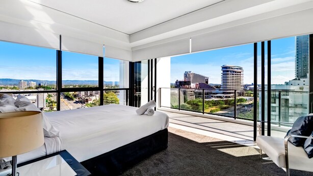 Una habitación de hotel moderna con una cama grande, una silla y vistas de la ciudad a través de amplios ventanales.