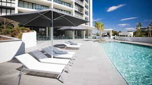 Zona de piscina moderna con tumbonas y sombrillas, rodeada por un edificio blanco con balcones.