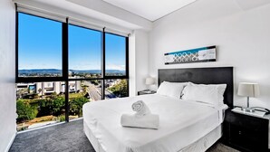 Una habitación de hotel moderna con una cama grande, dos mesitas de noche, vistas a un paisaje urbano a través de ventanales de piso a techo y una obra de arte abstracta en la pared.