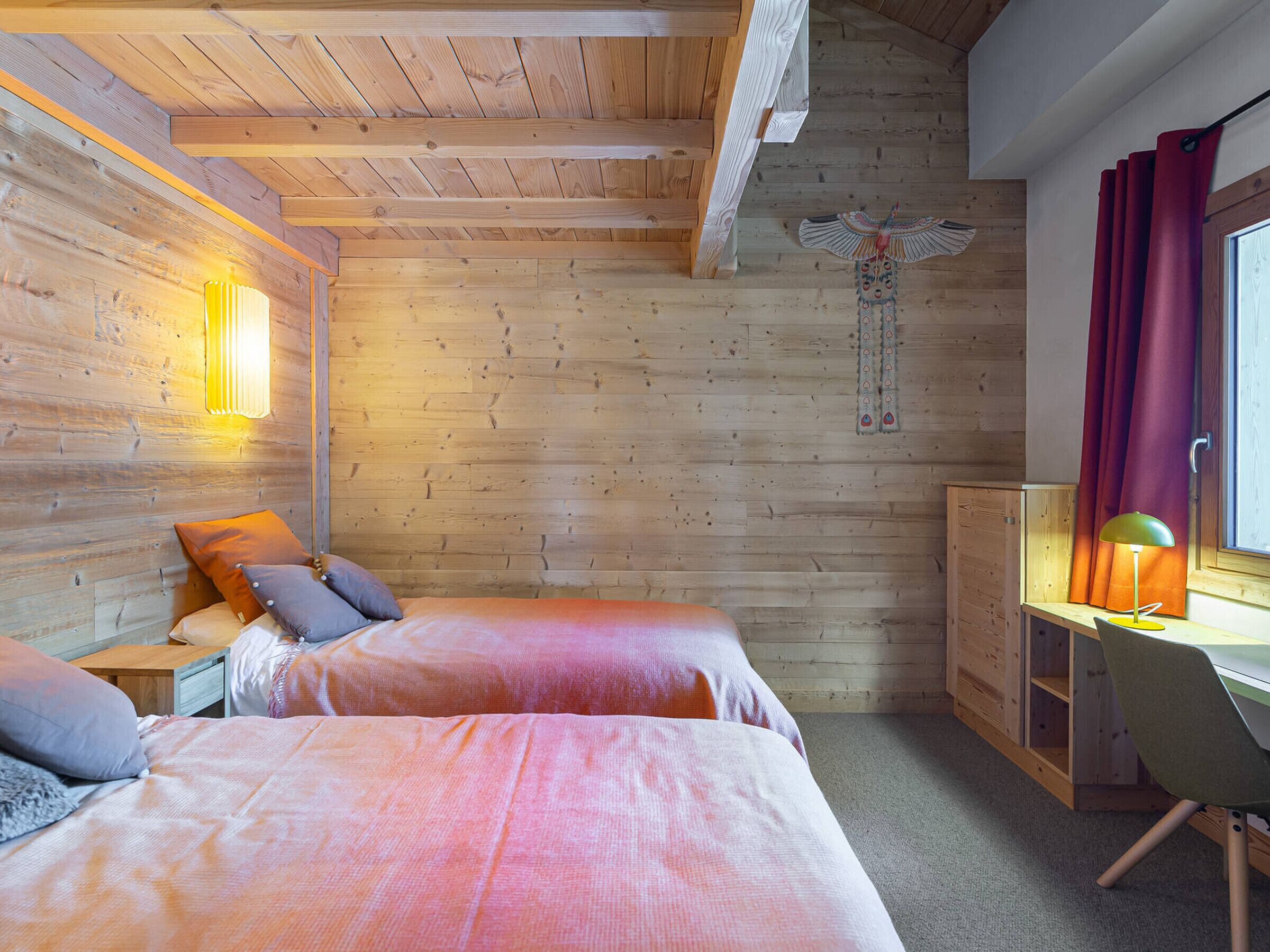 A wooden cabin room with two beds, a desk, a chair, and a window with curtains.
