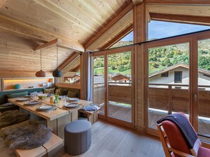 Ein Holzinnenraum mit Essbereich, einer Bank und Blick auf Häuser durch große Fenster.