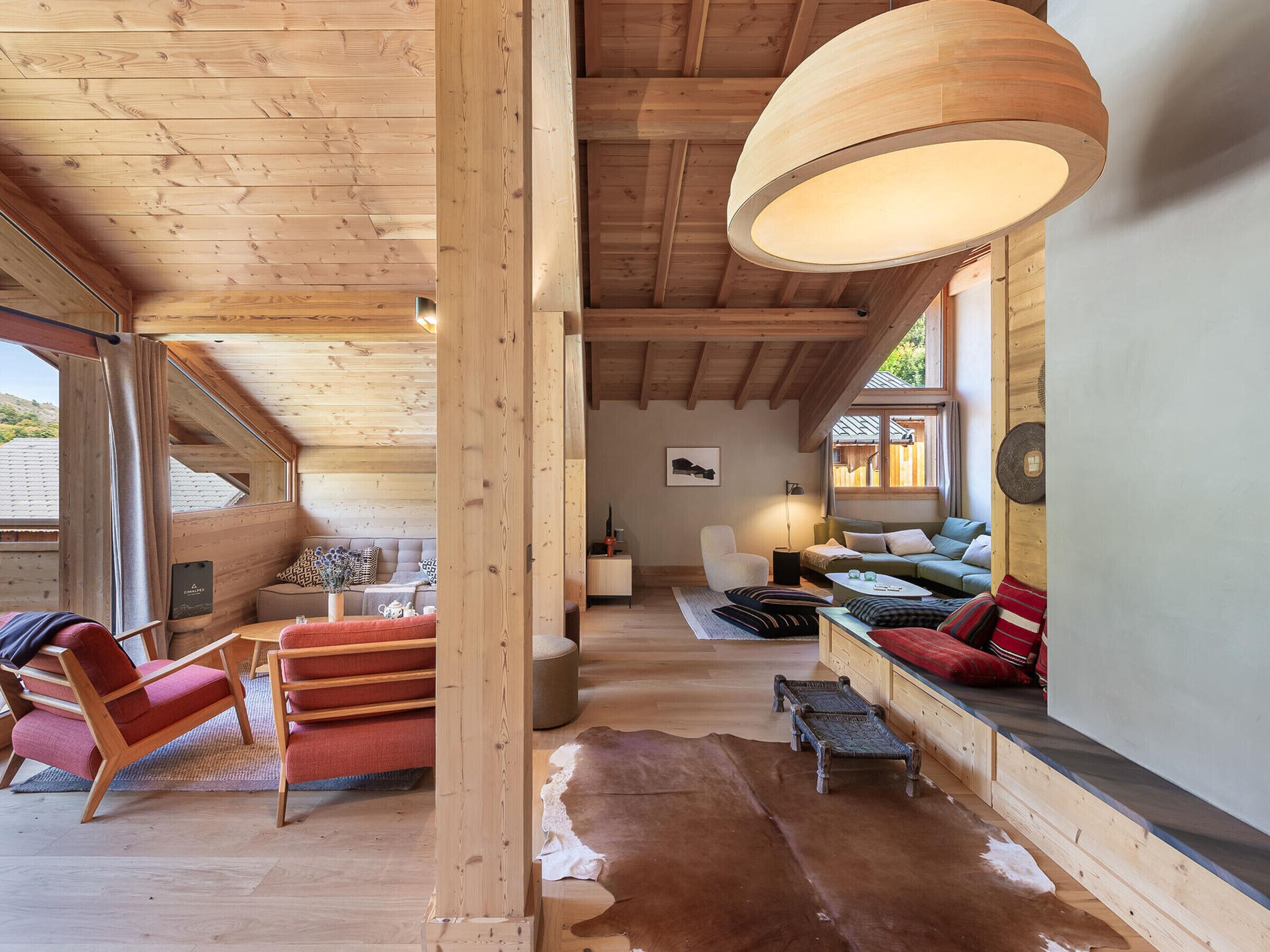 A cozy wooden interior with a high ceiling, a large wooden beam, a sofa, a chair, and a hanging light fixture.