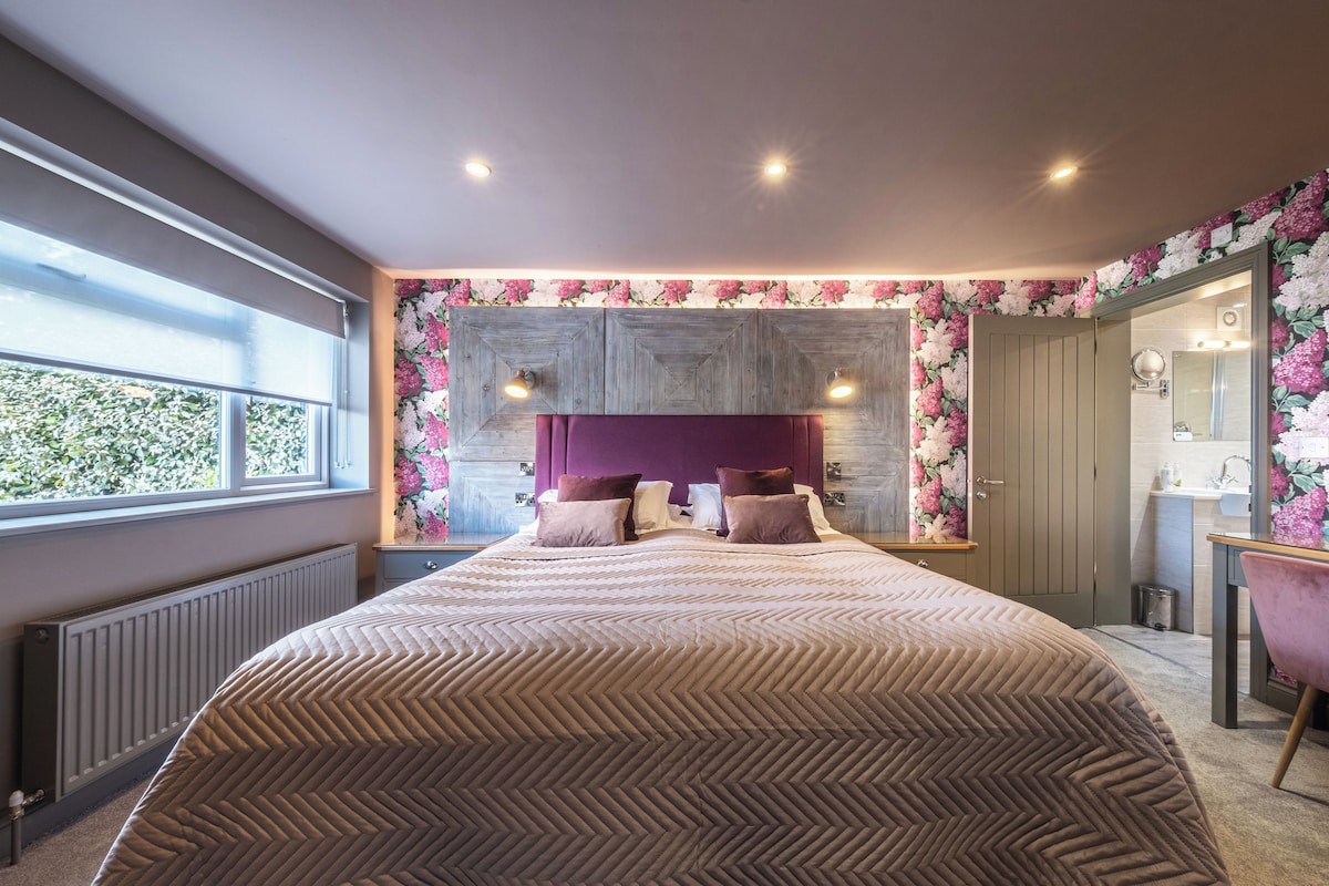 A bedroom with a large bed, a window, a bathroom with a mirror, and a floral wallpaper design.