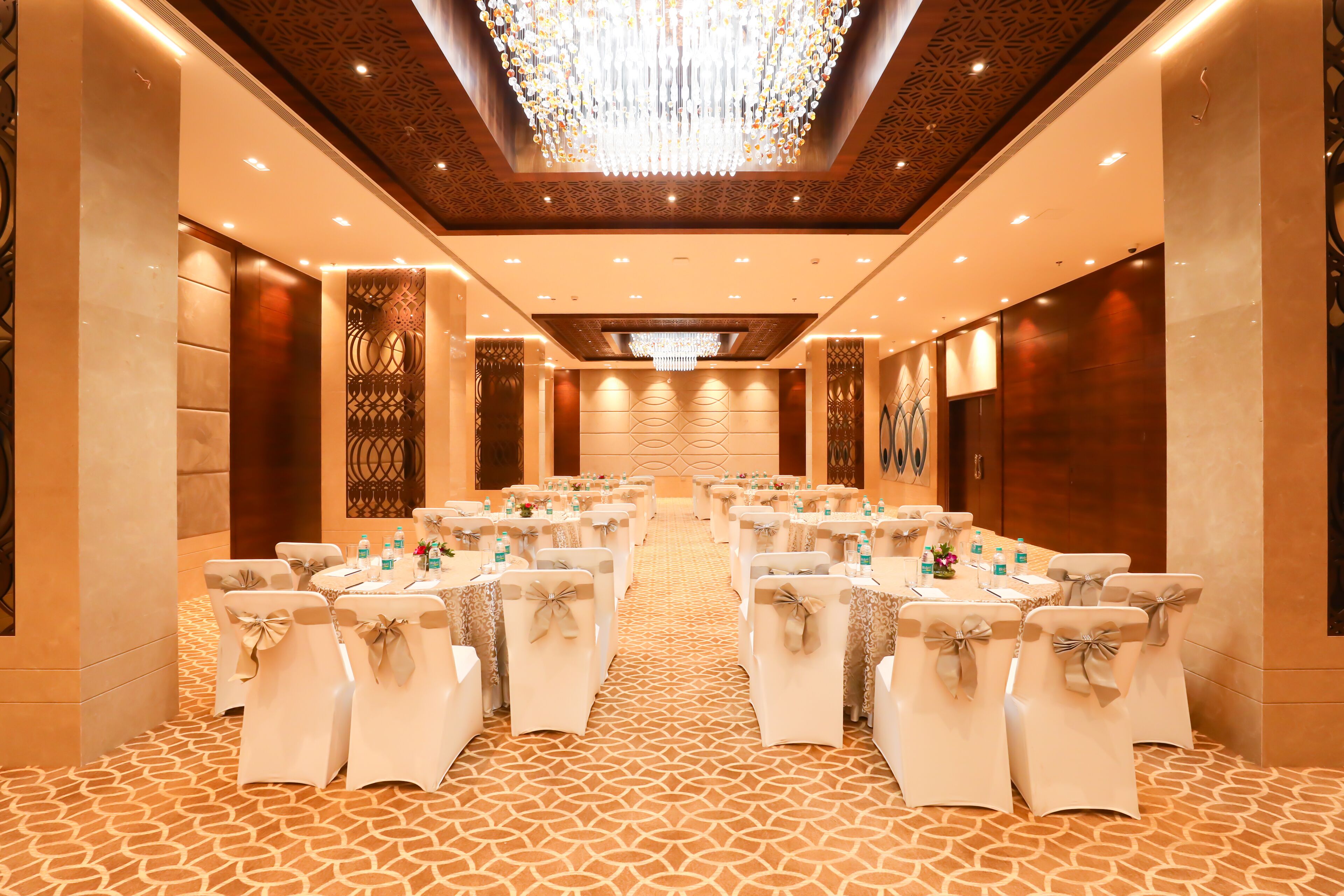A conference room with a patterned carpet, white chairs with bows, tables with water bottles, and a large chandelier.