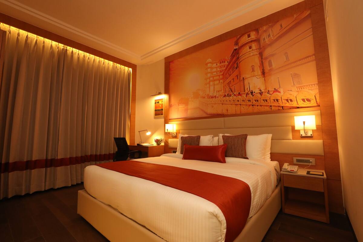 A hotel room with a large bed, a nightstand with a lamp, a wall-mounted phone, and a painting of a building.
