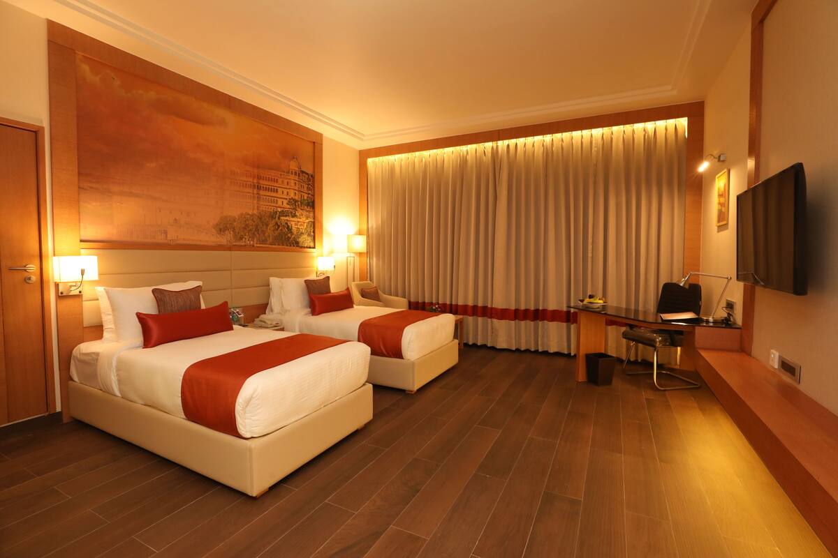 A hotel room with two beds, a desk, a chair, a television, and a large painting on the wall.