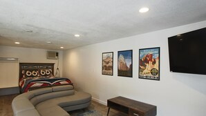 Un salon avec un canapé gris aux lignes courbes, une petite table d’appoint en bois et trois affiches encadrées accrochées au mur.