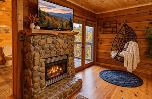 A stone fireplace with a fire, a hanging chair, and a view of autumn foliage through a window. - 10 min to Soaky mntns & shoping, private backyard (Sevierville)