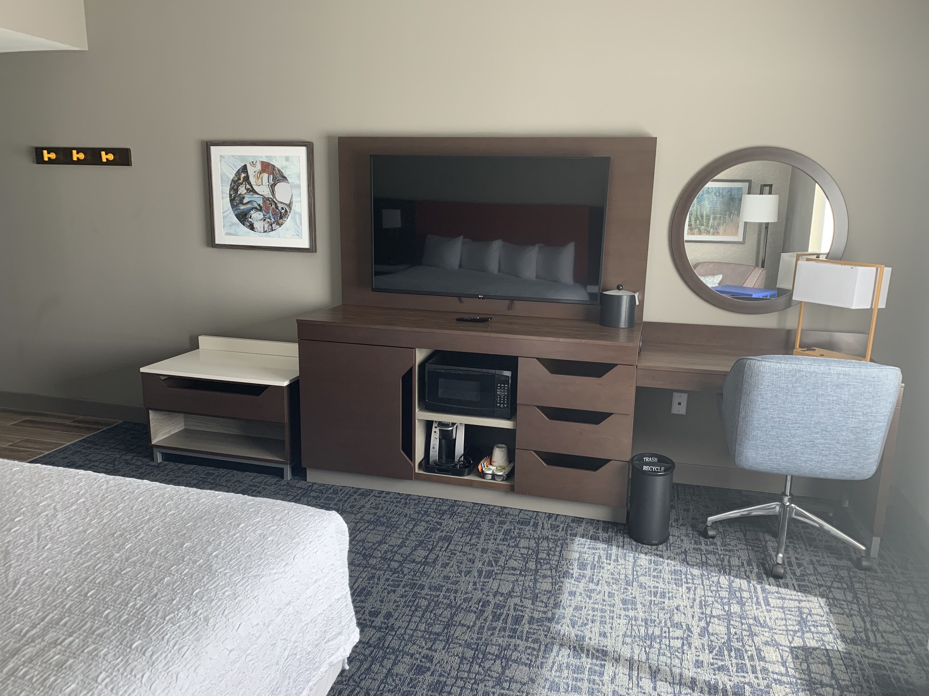 A hotel room with a bed, a desk with a television, a chair, a trash can, and a recycling bin.