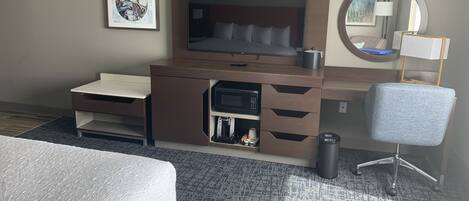 A hotel room with a bed, a desk with a television, a chair, a trash can, and a recycling bin.