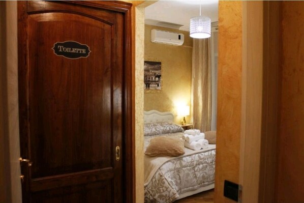 Ein Hotelzimmer mit einer Holztür mit der Aufschrift "TOILETTE", einem Bett mit gemusterten Bettbezügen, einer Nachttischlampe und einer Klimaanlage an der Wand.