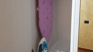 A closet with a hanging rack, an ironing board, and a shelf with an iron and a power strip. - St. Peter's Private Room (Roma)