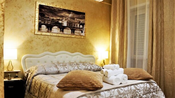 A neatly made bed with a patterned bedspread, two pillows, and a rolled-up towel. A framed picture hangs above the bed, and a nightstand with a lamp is visible to the left. - St. Peter's Private Room (Roma)