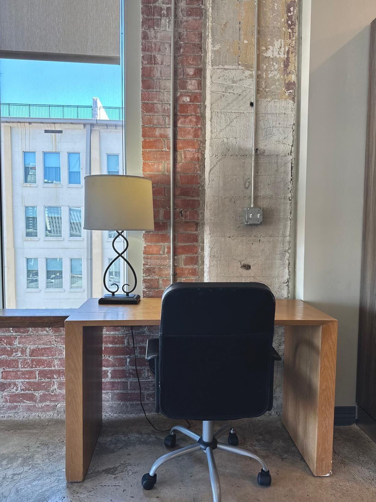 A corner office with a wooden desk, a black office chair, a lamp, and a view of buildings through a large window.