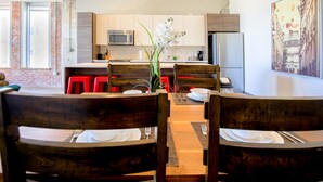 Una cocina moderna con armarios blancos, microondas y refrigerador. Hay una zona de comedor con sillas de madera y una mesa preparada para dos.