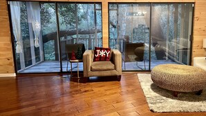 Ein gemütliches Wohnzimmer mit Holzboden, einem beigen Sofa mit Kissen 'JAY', einem Korbsessel und großen Glasüren, die auf eine Terrasse mit Blick auf Bäume führen.
