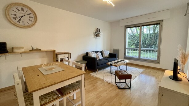 A modern living room with a wooden dining table, a black sofa, and a large window offering a view of greenery.