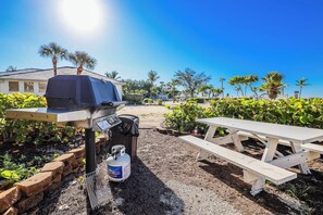 A barbecue grill with a propane tank, a picnic table, and a well-maintained garden with palm trees and shrubs. - Kimball Lodge #244 - Charming 1brm, 1 and a half bath, Beachside condo! (Sanibel)