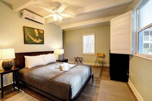A bedroom with a bed, a ceiling fan, an air conditioner, a window with blinds, a painting on the wall, and a small table with a lamp. - Kimball Lodge #244 - Charming 1brm, 1 and a half bath, Beachside condo! (Sanibel)
